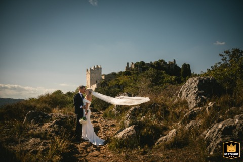 In Nice, France, a couple hugs as the sun sets, the bride's veil billowing in the wind. A distant castle on a hill adds to the romantic backdrop of their embrace.