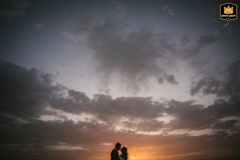 A couple is silhouetted against a sunset in Portugal. They pose low to the horizon with a big sky above, creating a dramatic and romantic portrait.