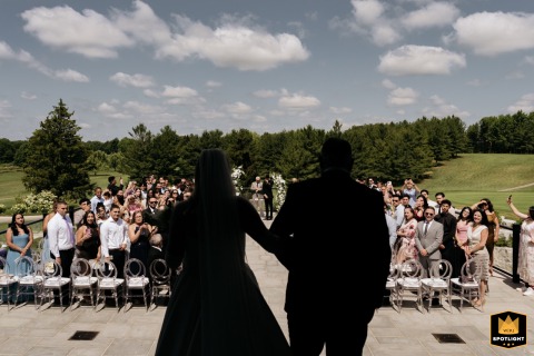 The bride makes her ceremony entrance in Toronto, silhouetted with her escort in shadow. Guests stand by their seats, bathed in sunlight in the background.