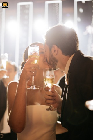 At a reception venue in Sao Paulo, the bridal couple shares a kiss during a toast. The intimate moment captures their joy amidst the wedding celebration.