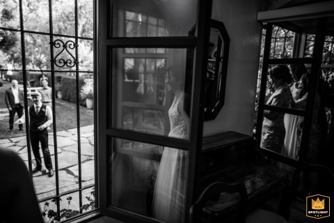 At Allied Arts in California, a bride playfully hides in anticipation during traditional wedding games. This black and white, layered photograph uses glass reflections to create a dreamy, artful composition, blending excitement, mystery, and joyful moment