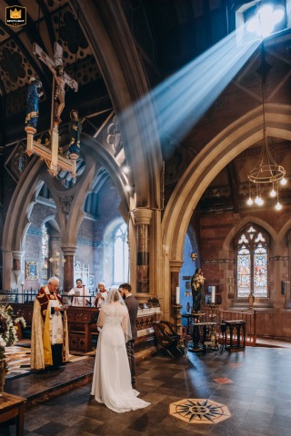 At All Saints Church in Babbacombe, Torquay, a beam of light dramatically illuminates the couple as they exchange vows beneath soaring gothic arches. The ethereal scene highlights romance, devotion, and the spiritual significance of their union.