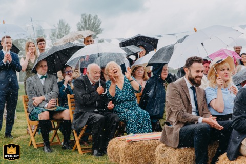 At Splotts Moor Farm in Glastonbury, guests laugh and cheer under colorful umbrellas as rain pours down during the open-air wedding ceremony. The joyful energy and shared resilience create unforgettable memories despite the unpredictable English weather.