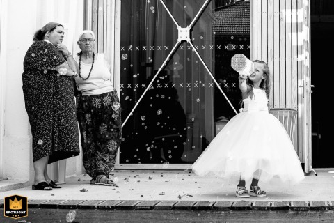 A candid, black-and-white photograph shows a little girl playing with a bubble machine. Older women talk nearby, creating a spontaneous wedding scene in Saint-Jean-de-Moirans, France.