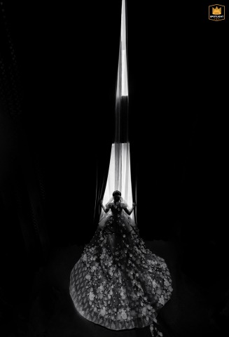 A black-and-white, vertical shot captures a bride looking out from a floor-to-ceiling window in Shenzhen, China. The tall composition emphasizes her pensive moment and the scale of the city.
