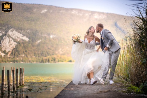 A bride and groom walk together along the water in Lépin-le-Lac, France. The couple shares a passionate kiss, capturing a romantic and intimate session during their wedding day.