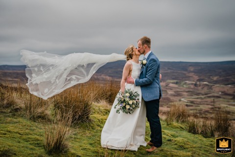 At The Priests House in Barden, Yorkshire, UK, the wedding couple poses in the breathtaking countryside. The bride’s flowing veil is caught by the wind, creating a romantic and timeless scene.