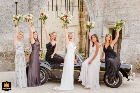 At Chateau de Lisse in Nerac, France, the bride and her bridesmaids stand proudly beside a vintage convertible sports car. With bouquets held high overhead, they celebrate together, radiating joy and excitement on this wedding day.