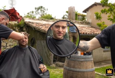 At The Castelnau in Lectoure, France, the groom enjoys an outdoor barber shop experience on the grass. A man holds a handheld round mirror, capturing a playful, candid wedding preparation moment in the fresh countryside air.