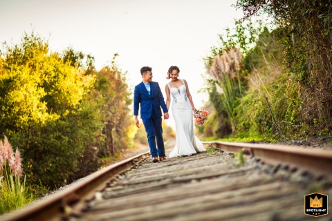 A newly married couple walks hand-in-hand down the railroad tracks in Capitola, California. The bride and groom pose for a portrait, creating a unique and romantic wedding day photograph.