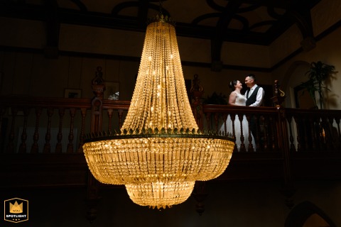 The bride and groom pose together for a photo indoors at Pendley Manor. They are positioned near a grand, ornate chandelier, which adds a touch of elegance and drama to the wedding portrait.