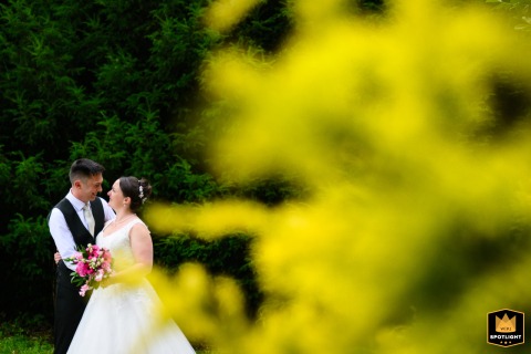 The bride and groom pose for a portrait at Pendley Manor. Soft-focus light green foliage in the foreground frames the couple, creating a romantic and natural-looking wedding photograph.