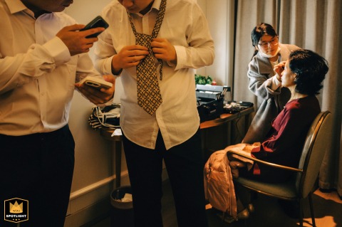 A Wedding Morning in Kaifeng: A Mother's Touch and a Groom's Friends Groom and friends learning to tie a tie while mother applies makeup in Kaifeng, China.