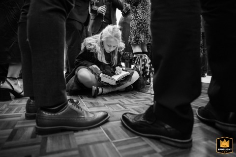 Marquee Marvels: A Young Girl's Quiet Corner Amidst the Wedding's Feet Young girl in a marquee at a private home, reading a book, surrounded by wedding guests' feet.