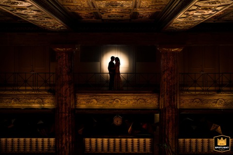 Love’s Center Stage: A Spotlight Shines on the Newlywed Couple in Philadelphia Philadelphia, Pa. — A wedding photojournalist captures a couple’s love taking center stage at the Ballroom at the Ben, silhouetted by a spotlight against a backdrop of golden columns.