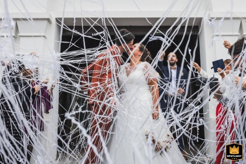 Exiting Ceremony, a Shower of Confetti for the Newlywed Couple A documentary-style photograph captures a joyful moment as a newlywed couple exits their religious ceremony in the Basque Country, France. The image shows the bride and groom surrounded by an explosion of confetti and white ribbons.