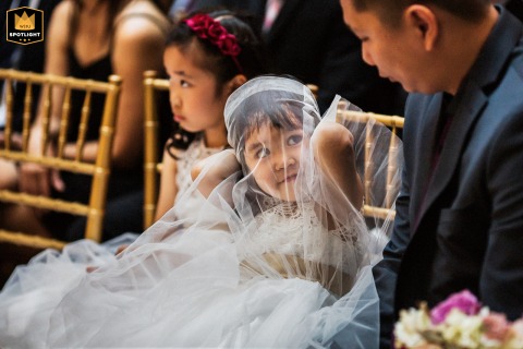 Documentary Glimpse: Two Girls and a Veil at a San Francisco Wedding A documentary-style photograph captures a moment during a wedding ceremony at the San Francisco Design Center. The image shows two girls sitting in chairs; one is under a tulle veil and is looking at the guest next to her.