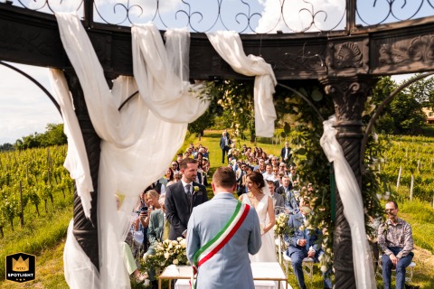 Symmetrical Scene of a Sunlit Italian Wedding Through an Arbor A documentary-style photograph captures a symmetrical moment from behind the officiant at a ceremony in San Floriano del Collio, Gorizia, Italy. The image is shot through an arbor, highlighting the sunlit outdoor setting.