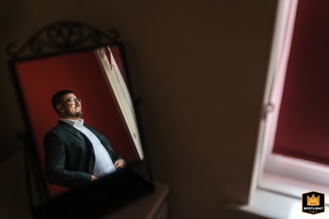 Reflections Of A London Groom's Calm Preparations In A Mirror A wedding photographer captures a serene scene at a London hotel. The image, framed by a mirror, shows the groom getting ready by a window, bathed in soft, natural light.