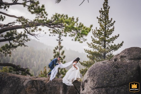 Lake Tahoe Nuptials: Walking Amidst Boulders Under Pines, Serene and Foggy In South Lake Tahoe, California, a wedding photographer captures a serene image on a foggy day. The image shows the couple carefully walking among boulders under pine branches.