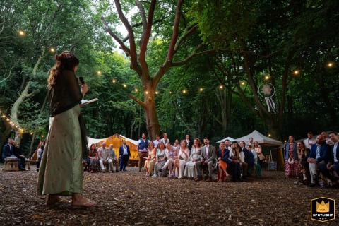 Heartfelt Vows and Golden Light Under the Woodland Canopy A wedding photographer captures heartfelt speeches under a canopy of trees at Woodland Weddings. The enchanting image shows guests listening intently, illuminated by warm strings of lights.