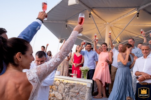 Raising a Toast to Love at Sunset in Symi, Greece At the Sunset Restaurant in Symi, Greece, a WPJA wedding photographer captures a joyous moment. All wedding guests raise their glasses in a toast, enthusiastically celebrating and greeting the newly married couple.