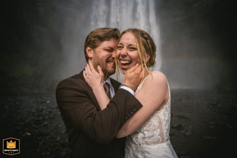 Bride And Groom Laugh Joyfully Before Majestic Skogafoss Waterfall In Iceland Portrait Skogafoss, Iceland: A joyful portrait of a bride and groom laughing heartily with the magnificent waterfall as their backdrop.