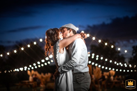 Maine-et-Loire Wedding Kiss Beneath A Canopy Of String Lights Maine et Loire, France: Couple shares a kiss at night, string lights stretching across the frame behind them.