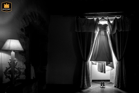 Bridal Gown Dramatically Lit by Sun and Lamp at Château des Girards Château des Girards, France, a bridal gown hangs dramatically in a brightly lit window, its edges glowing from the sun, while a small, glowing lamp offers a contrasting warm light across the frame.