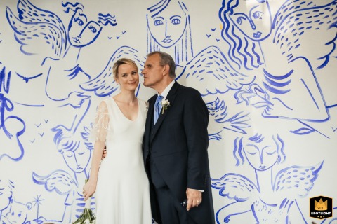 Couple Poses Against Ornate Angel Wall Inside Historic Auch Chapel In Auch, France, a newly married bride and groom pose inside a historic chapel, standing close together against a striking and ornate wall decorated with printed angels rendered dramatically in blue ink.