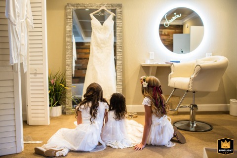 Three Young Bridal Party Members Intimately Gaze at the Hanging Wedding Dress At the Deer Park Villa in Fairfax, CA, three young bridal party members in their wedding attire sit closely together, intimately gazing at the bride's wedding dress as it hangs nearby before the ceremony begins.