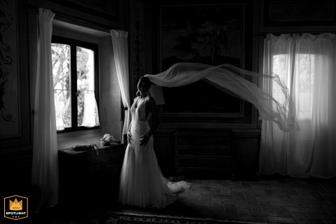 Black and white portrait of a bride indoors by a window, with her veil flowing in the air at Palazzo Viviani, Monte Gridolfo, Rimini, Italy.