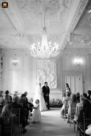 Black and white vertical photo at Longstowe Hall in Cambridge, UK, featuring a flower girl laughing with her dad during the ceremony, framed by the venue’s striking architecture.