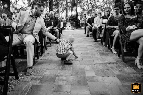At Deer Park Villa in Fairfax, California, a man seated in the front row gently reaches out to contain a toddler standing alone in the middle of the wedding aisle, captured in black and white.