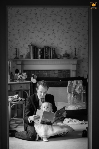 At a private home in Northamptonshire, East Midlands, a black-and-white photo captures a seated usher reading a book to the flower girl, highlighting a sweet and peaceful connection between generations amid the wedding festivities.