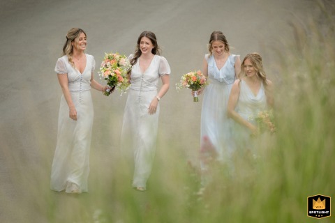 At St Peter & St Paul Church, Wing, Rutland, bridesmaids walk together along a dirt path bordered by tall green grass blades, the natural setting lending a fresh, serene backdrop to their procession.