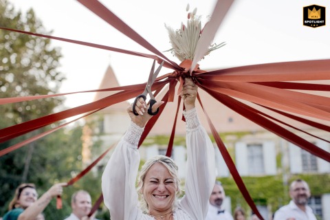 At Château de La Salle, la Pacaudière, the bride stands outside with eyes closed, holding scissors overhead to cut ribbons during the bouquet game, surrounded by eager guests hoping to win the prize.