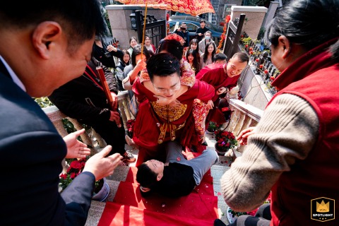 In Taiyuan, Shanxi, the groom carries the bride on his back up red-carpeted stairs, surrounded by smiling family. A playful boy stumbles ahead of them, adding a lively touch to the joyful scene.