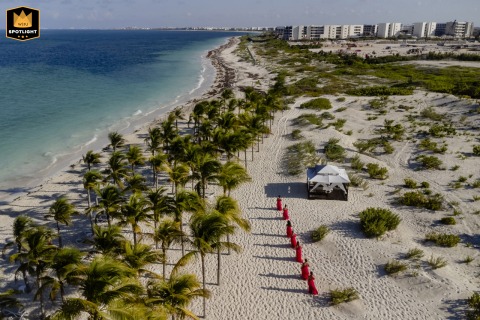 Aerial drone view of Puerto Cancun shows the bridal party in matching red dresses walking in a line across sandy beach, their vibrant attire creating a striking contrast against the natural seaside backdrop.