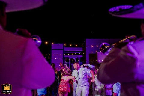 A joyful couple in Tulum link arms and swing each other in a lively circle on the dance floor, surrounded by festive lighting and laughter, their expressions filled with happiness and celebration.