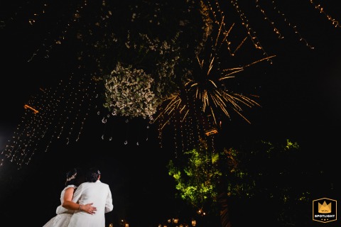 The bride and groom stand close together on the beach at night, viewed from behind as vibrant fireworks burst overhead, illuminating the Tulum sky and casting a glow over their celebration.