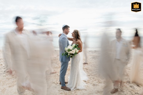 A stunning long exposure captures the bride and groom standing on soft Tulum sand while their bridal party moves in a gentle circle around them, creating dreamy pastel light trails and a romantic, ethereal feel.
