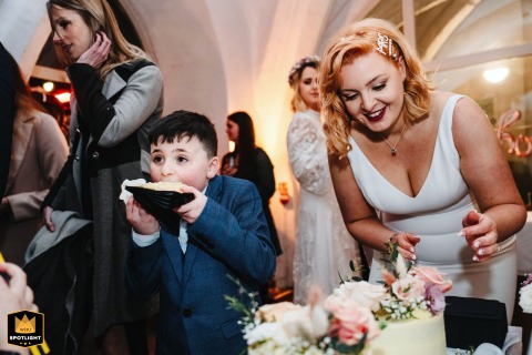 At Cinema City in Norwich, UK, a delighted young boy eagerly leans over the cake table beside the bride, ready to stuff a piece of wedding cake into his mouth, capturing pure childhood excitement.