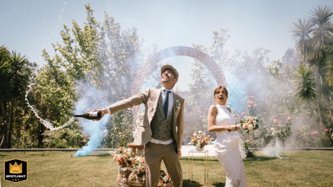 A joyful bride and groom arrive at Quinta Largo da Vila in SJMadeira, Portugal, laughing together as they are surrounded by vibrant blue smoke from grenades. Champagne sprays in the air, setting a festive celebration atmosphere outside the beautiful venue