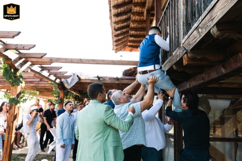 In Plovdiv, the groomsmen enthusiastically lift a man up to a wooden balcony as part of a spirited wedding tradition, infusing the celebration with an air of danger, excitement, and bold adventure against the backdrop of historic architecture.