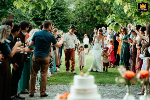 At Quinta da Eira in Penafiel, Portugal, the bride and groom walk with two children down a grassy aisle, framed by smiling guests outdoors, heading together toward the cake-cutting celebration under the canopy of trees.