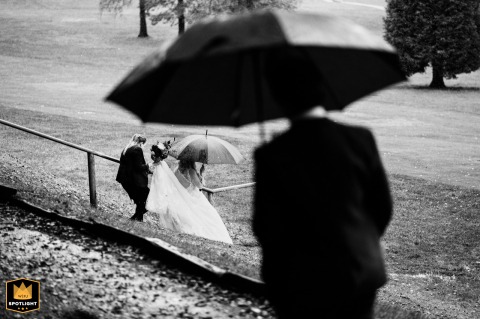 At Old Thorns Hotel in Hampshire, UK, during a classic rainy British wedding, the bride is captured in the background walking down steps on the golf course, perfectly framed by an umbrella, with vintage black and white tones highlighting the timelessness.