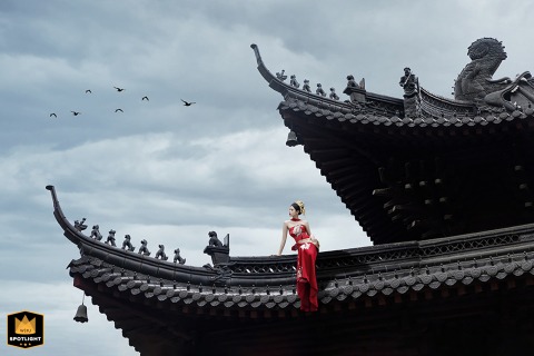 At a Chinese church, the bride waits for the groom while seated elegantly on a balcony in a red dress, looking contemplative as birds soar overhead, highlighting her anticipation against a graceful and serene backdrop.