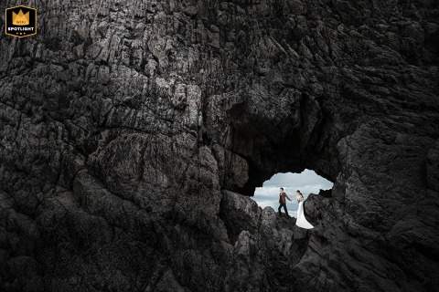 In Fujian, China, the bride and groom pose for a seaside portrait, framed through a natural opening in a rock formation resembling a cave, emphasizing the dramatic coastal landscape and their elegant presence against the ocean backdrop.