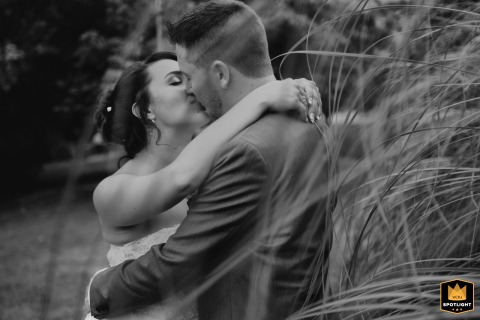 In the expansive park of Mondonville, Haute-Garonne, the bride and groom share a passionate kiss beside tall, wispy grasses, captured in black and white to highlight the romance and natural surroundings of their wedding day.